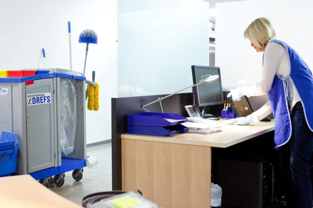 Ein Arbeiter reinigt einen Büroschreibtisch neben einem blauen Reinigungswagen und Computermonitoren.