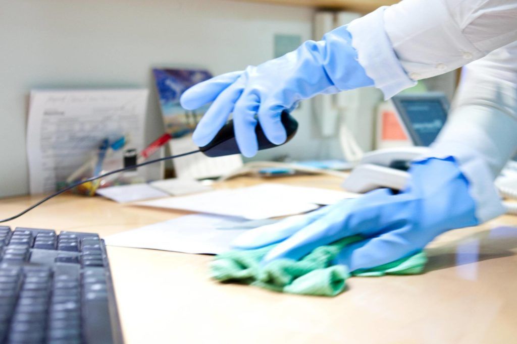 Behandschuhte Hände reinigen einen Schreibtisch mit einem blauen Tuch in der Nähe einer Tastatur und Büromaterialien.