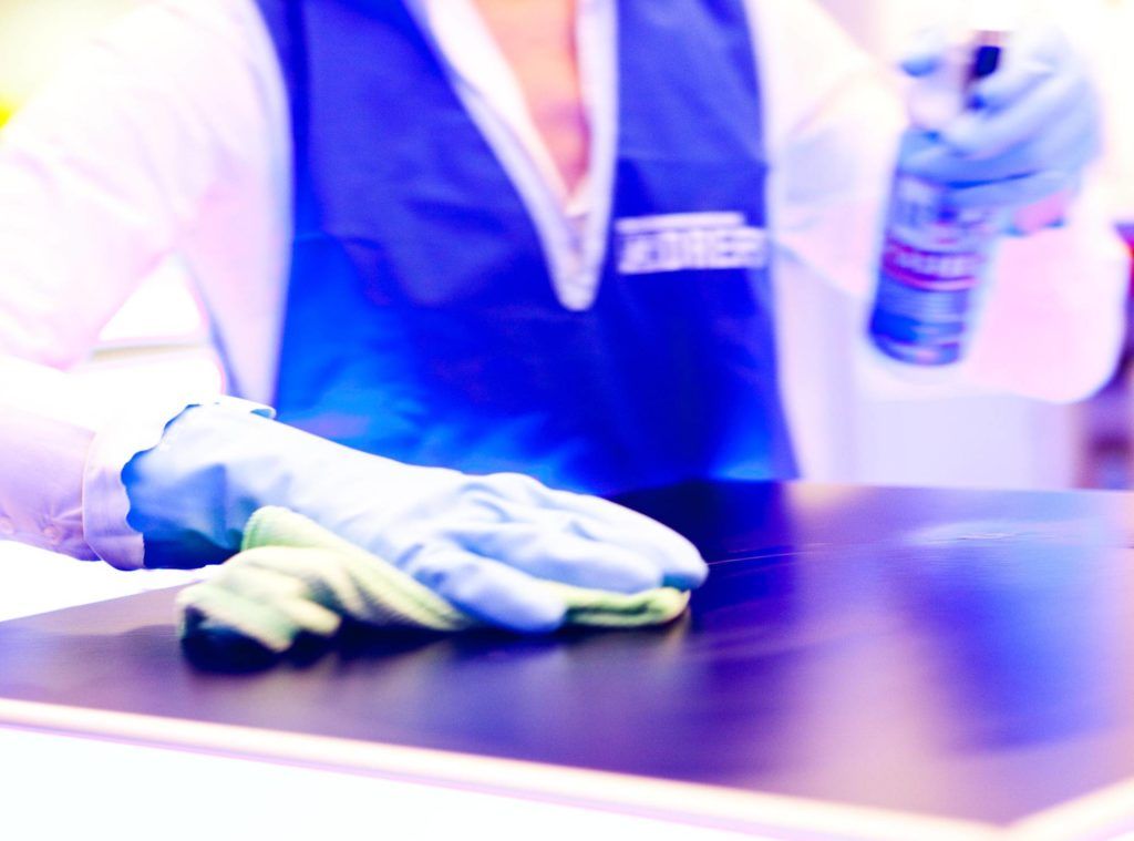Eine Person mit blauen Handschuhen reinigt in der Nähe eine dunkle Arbeitsfläche mit einer Sprühflasche.