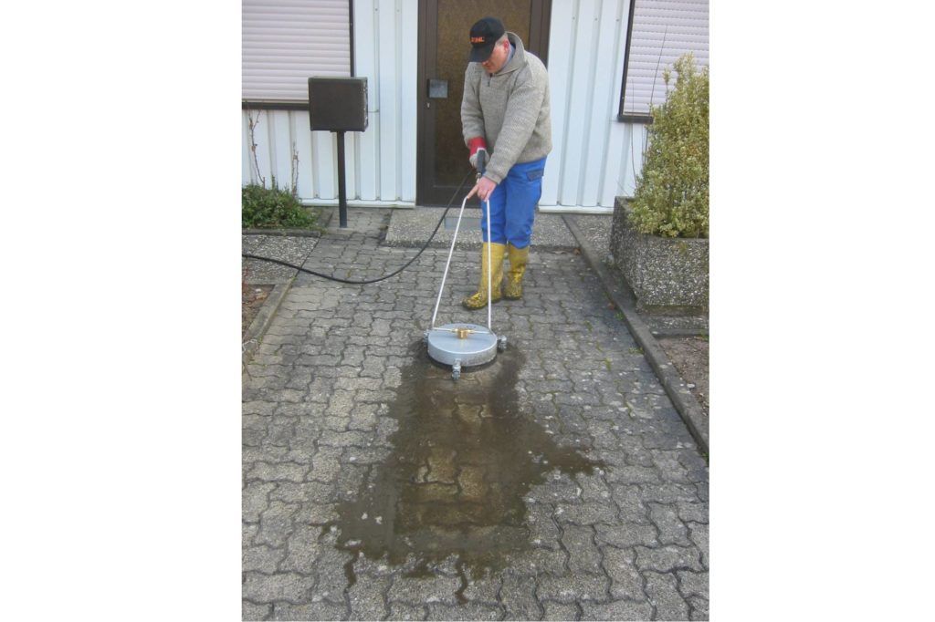 Eine Person reinigt eine nasse Ziegelsteinauffahrt vor einem Haus mit einem Hochdruckreiniger.