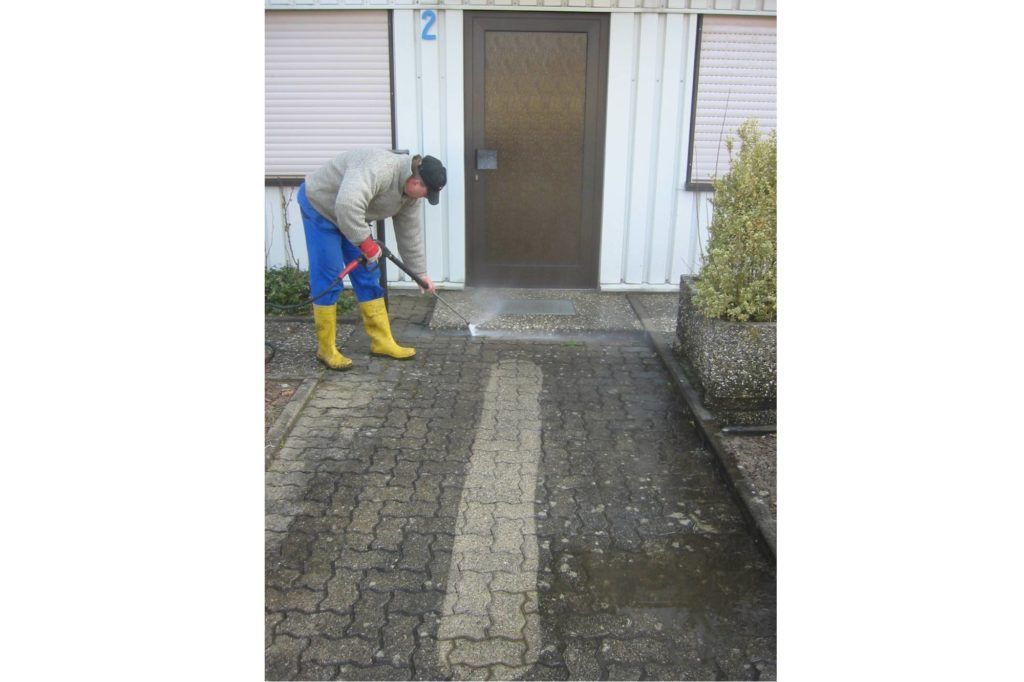 Eine Person reinigt mit einem Gartenschlauch eine Einfahrt in der Nähe eines Hauseingangs.