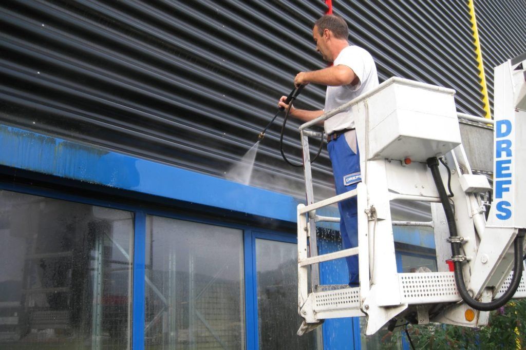 Ein Arbeiter reinigt auf einer Hebebühne die Außenfassade eines blauen Gebäudes mit einem Hochdruckreiniger.