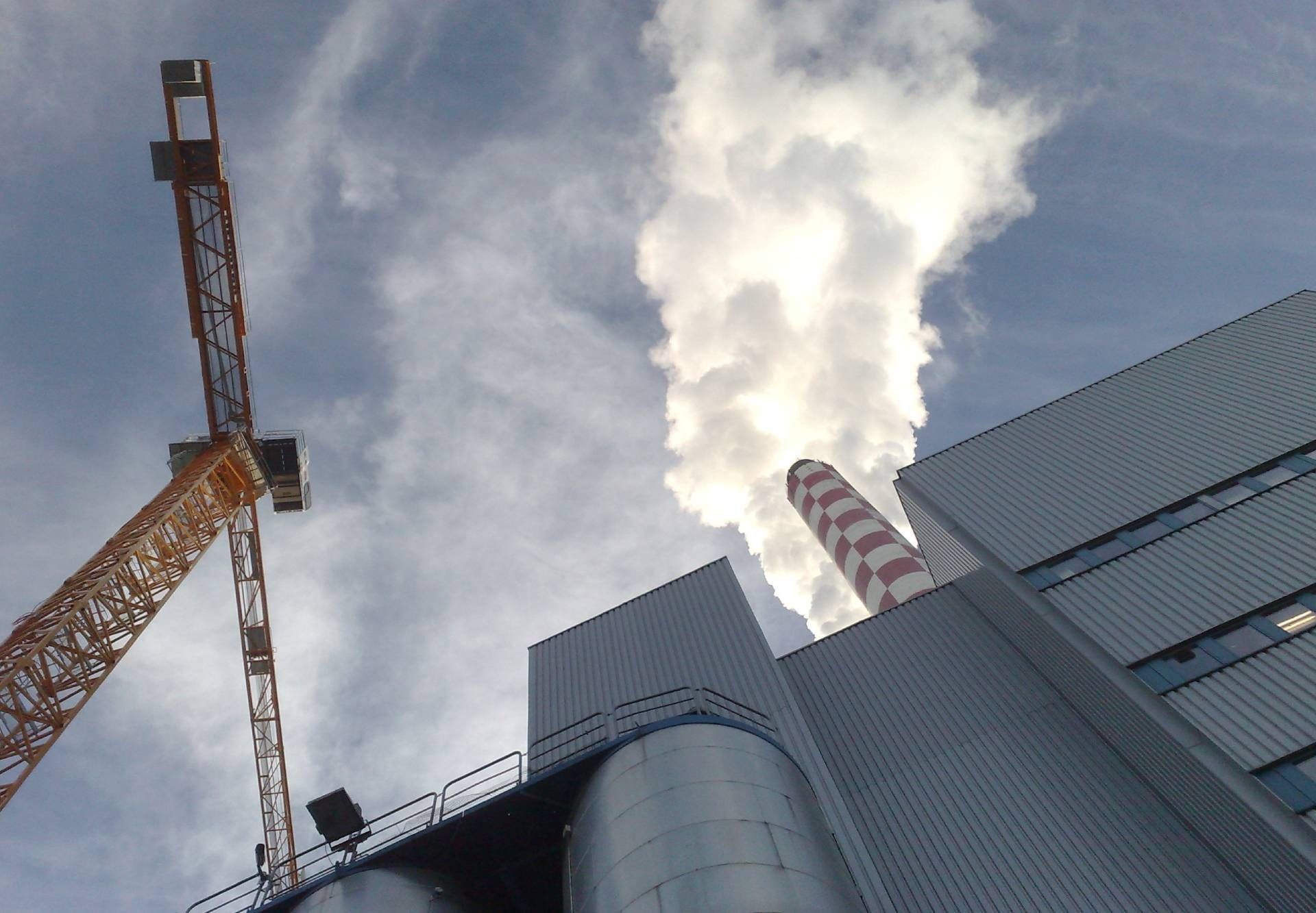 Industriegebäude mit Kränen und einem Schornstein, aus dem dicker, weißer Dampf in den Himmel steigt