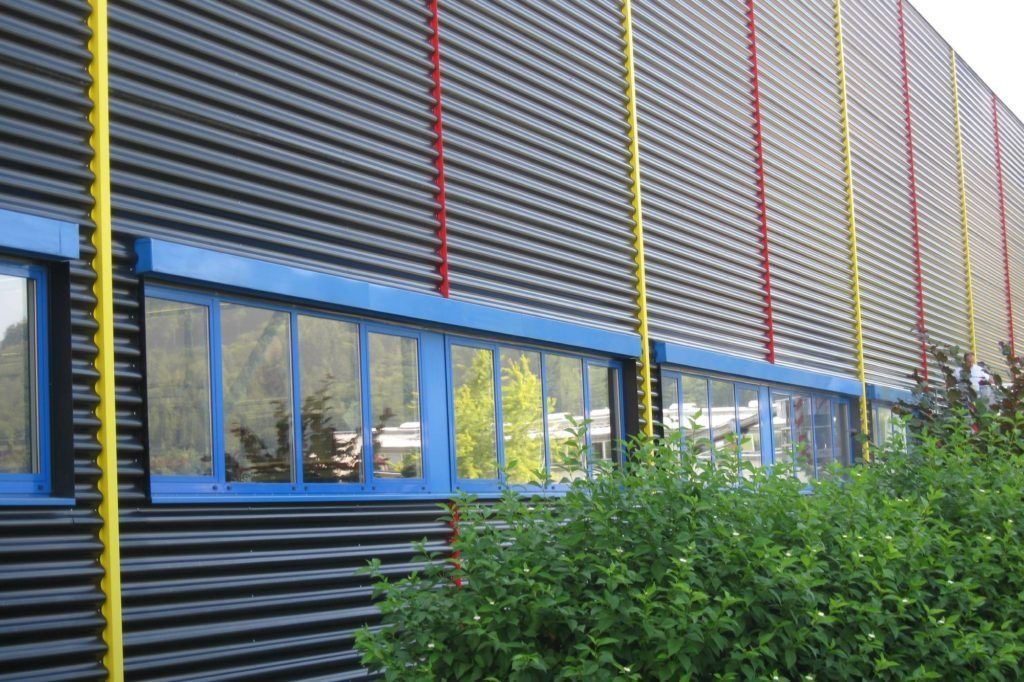 Moderne Gebäudefassade mit blau umrandeten Fenstern, grauer Metallverkleidung und Sträuchern davor