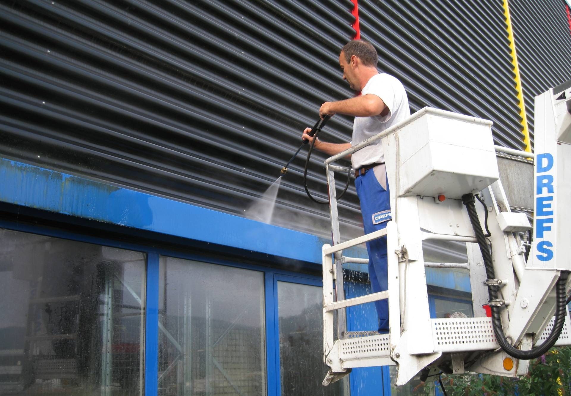 Ein Arbeiter in einer weißen Hebebühne reinigt mit einem Hochdruckreiniger ein dunkelblaues Schaufenster.
