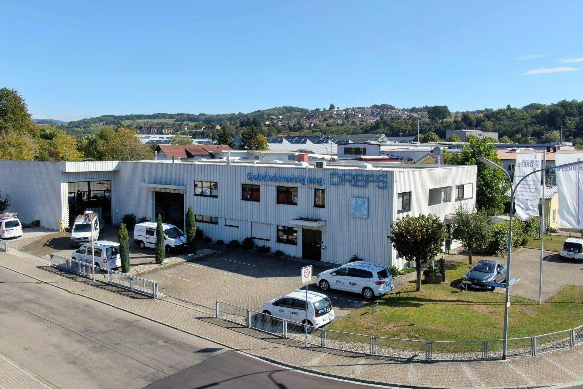 Weißes zweistöckiges Bürogebäude mit geparkten Autos und Werbebannern in einer sonnigen Vorstadtlage