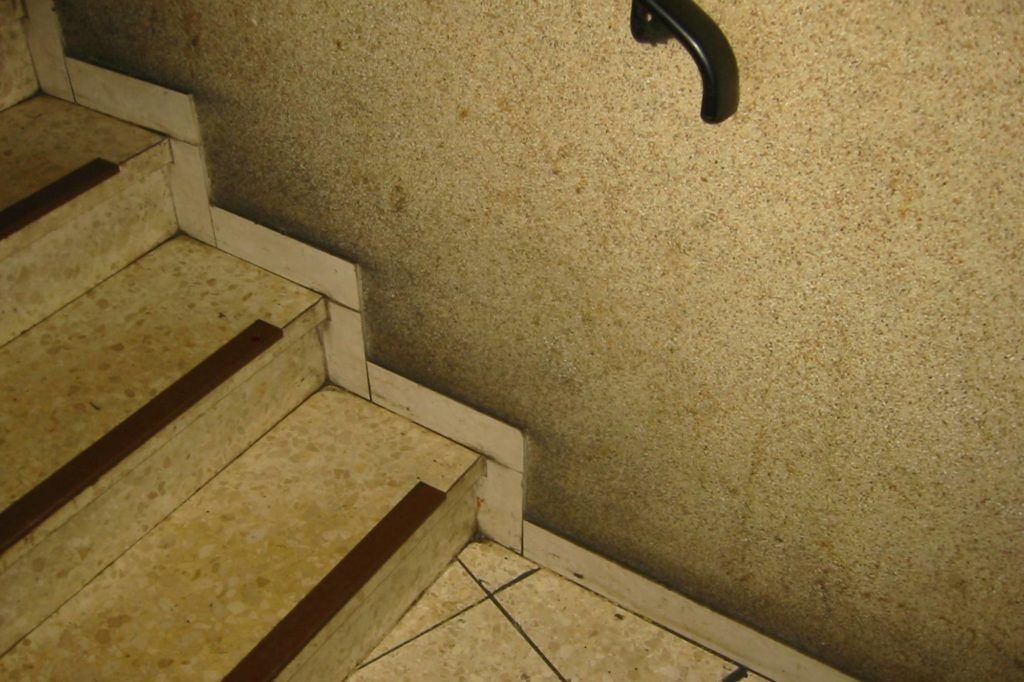 Treppenhaus mit beigefarbenen Fliesenstufen und schwarzem Handlauf an einer strukturierten Wand.