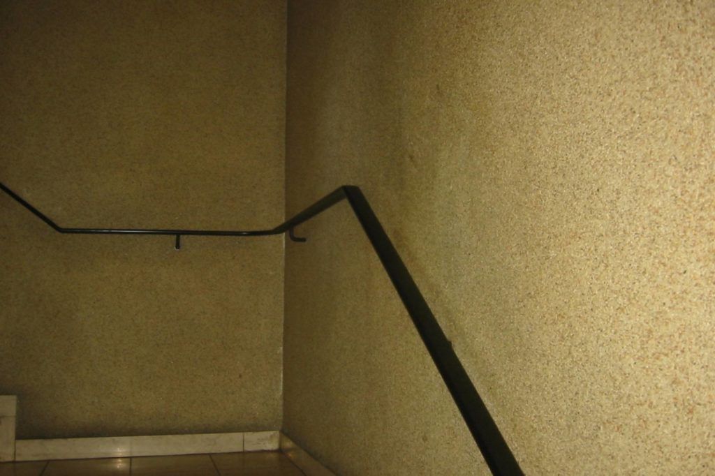 Dunkle Treppenhausecke mit beigefarbenen, strukturierten Wänden und schwarzem Handlauf.