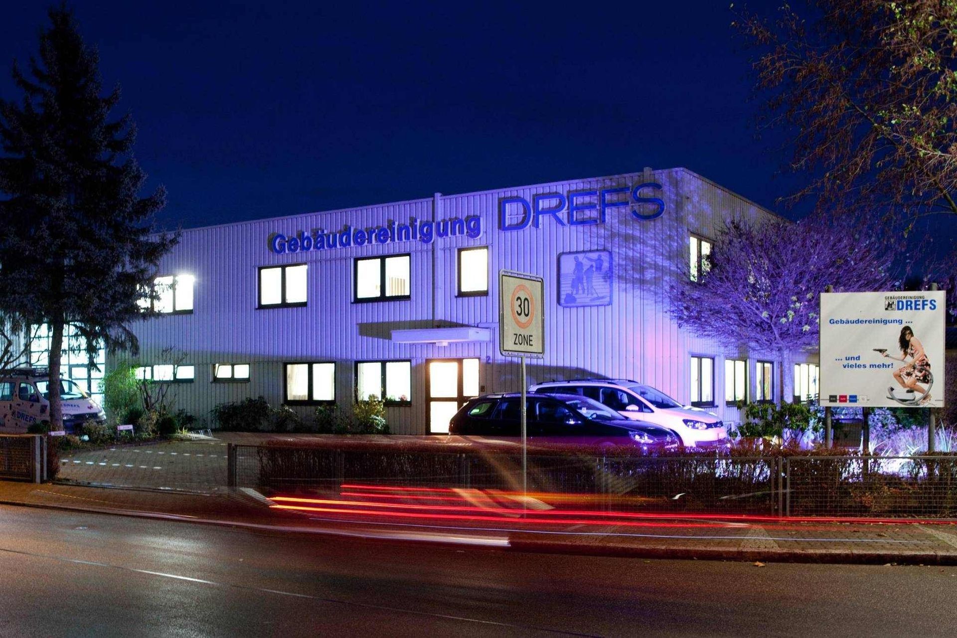 Nächtliche Straßenansicht eines beleuchteten Bürogebäudes mit blau-violetter Fassade und Scheinwerferlichtspuren davor