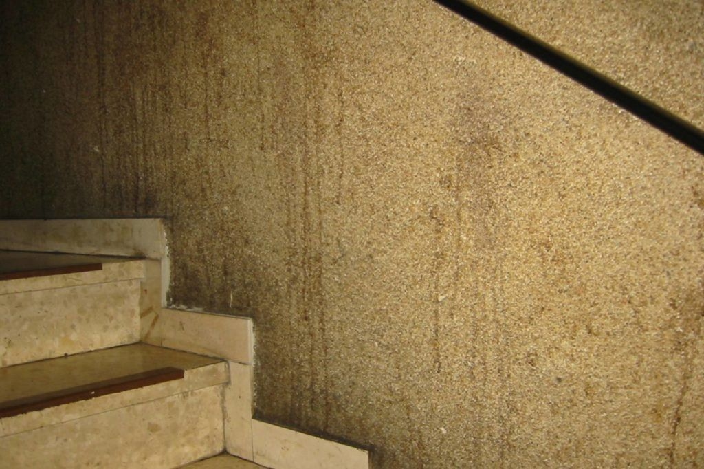Nahaufnahme einer beigefarbenen Steintreppe neben einer strukturierten, hellbraunen Wand mit schwarzem Handlauf.