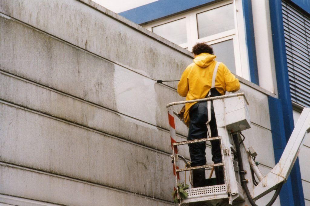Ein Arbeiter in einer gelben Jacke steht auf einer Hebebühne neben einer Gebäudewand und inspiziert die Außenfassade.