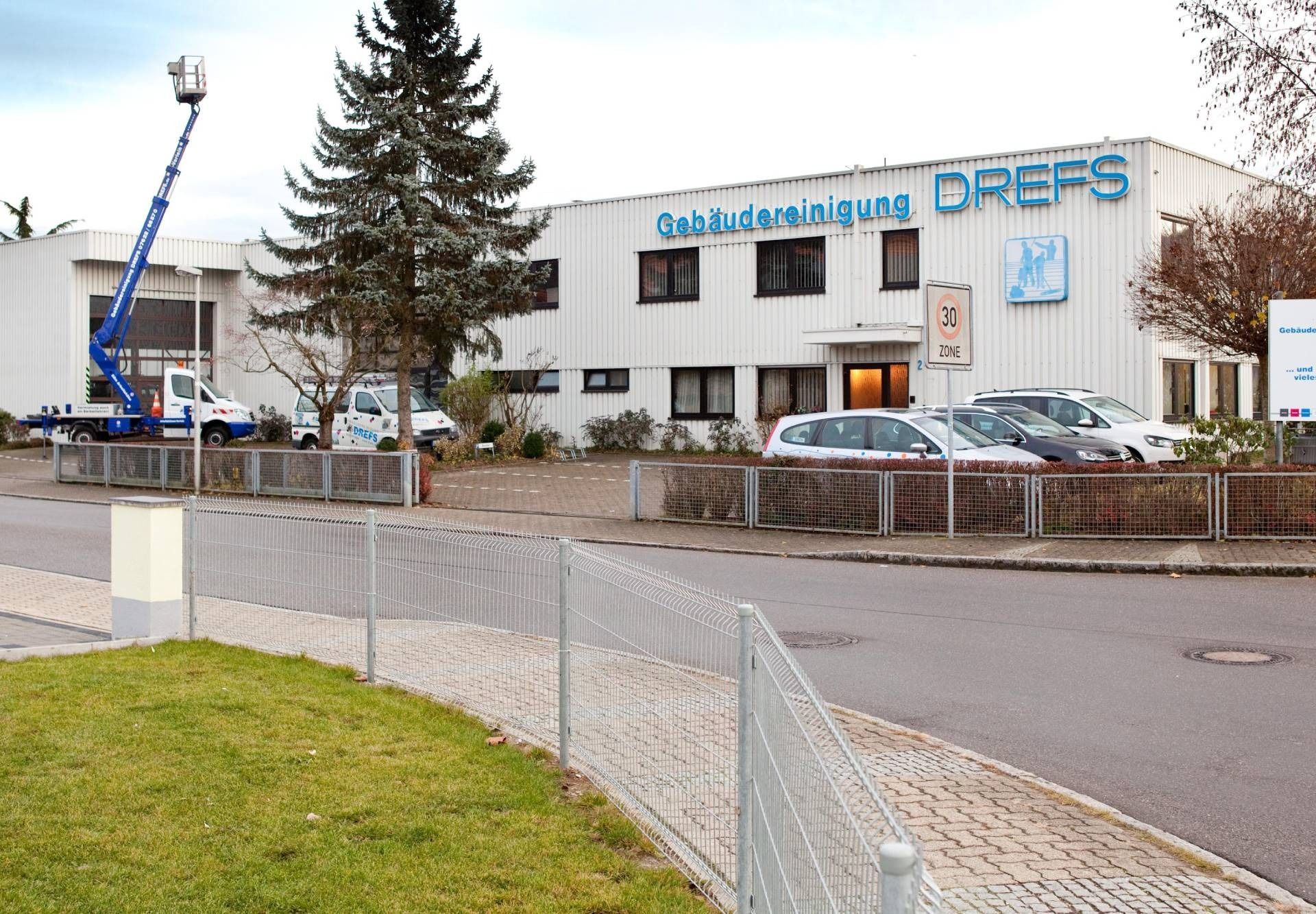 Straßenansicht eines weißen Gebäudes mit blauer Beschilderung, geparkten Autos und einer Hebebühne im Außenbereich.