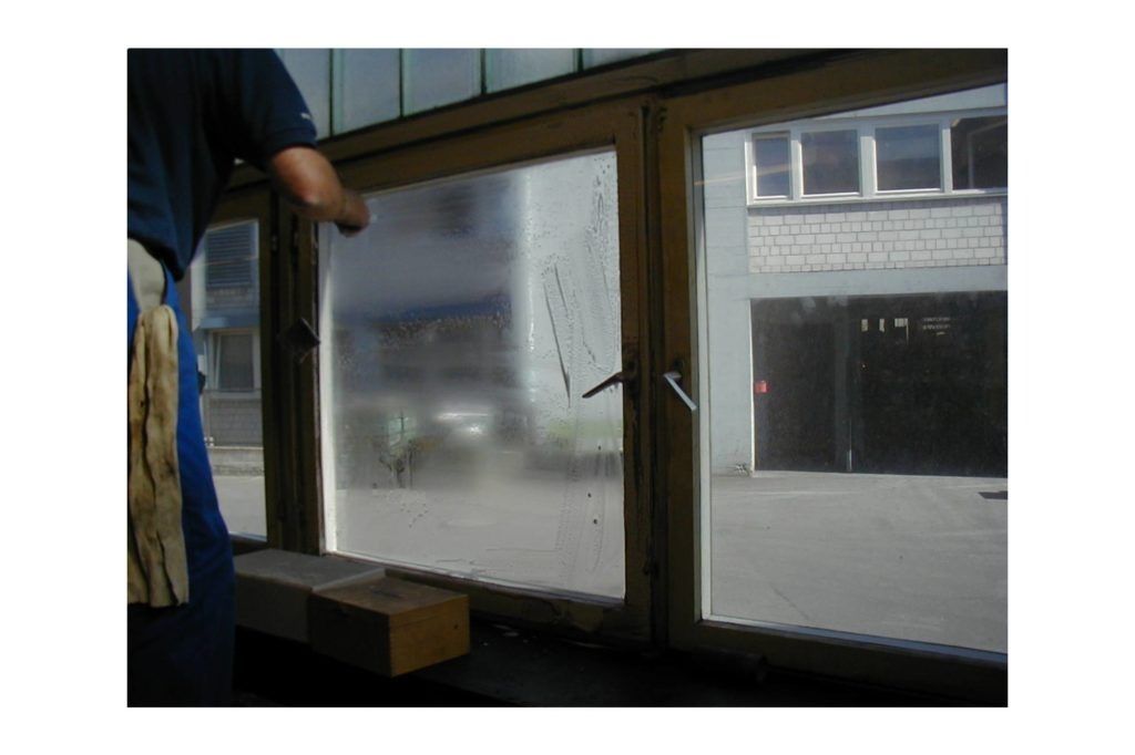 Eine Person putzt ein beschlagenes Fenster in einem Gebäude; draußen ist die Straße zu sehen.