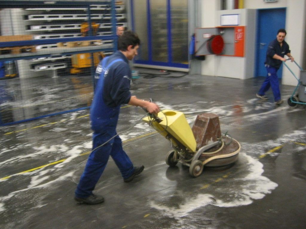 Arbeiter reinigen einen nassen Fabrikboden mit einer gelben Scheuersaugmaschine.