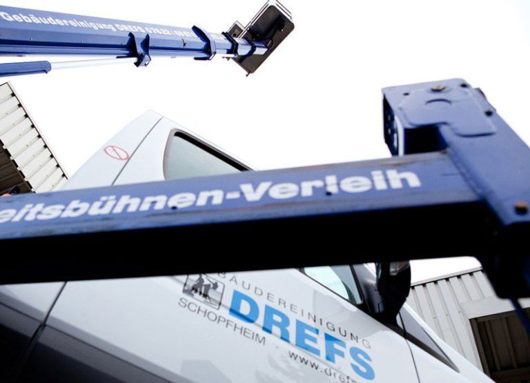 Ein blauer Kran hebt ein weißes Schild mit deutscher Aufschrift vor dem Himmel hoch.