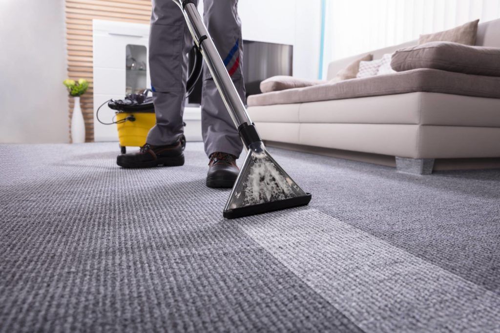 Eine Person saugt in einem hellen Wohnzimmer einen grauen Teppich; ein gelber Eimer steht daneben.