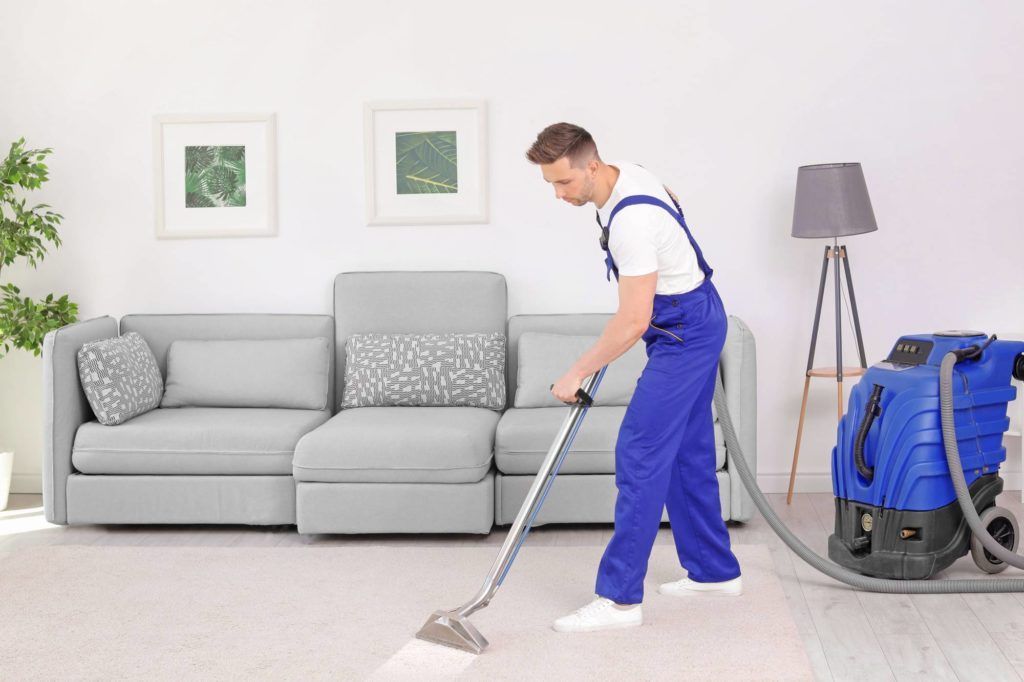 Eine Reinigungskraft saugt Staub in einem hellen Wohnzimmer neben einem grauen Sofa und einer Stehlampe.