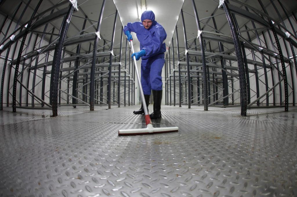 Eine Person in blauer Arbeitskleidung fegt einen Metallboden in einer großen Industriehalle.