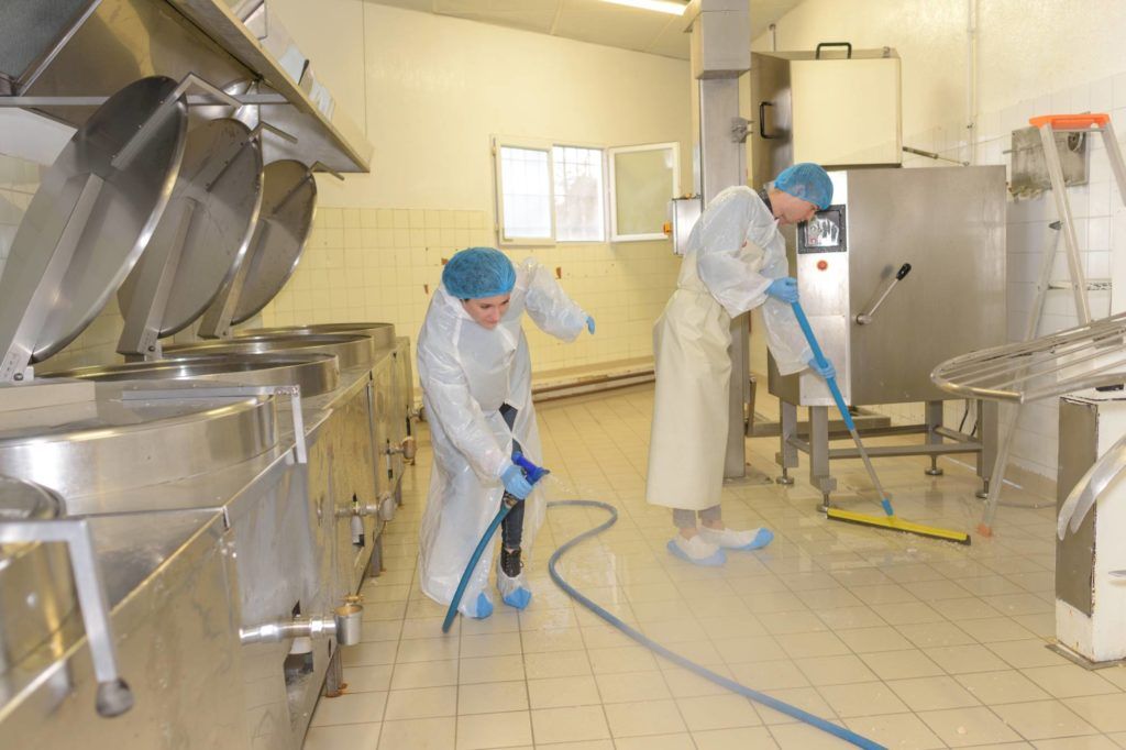 Arbeiter in weißen Kitteln und blauen Hauben reinigen mit Schläuchen und Werkzeugen den Edelstahlboden einer Fabrikküche.