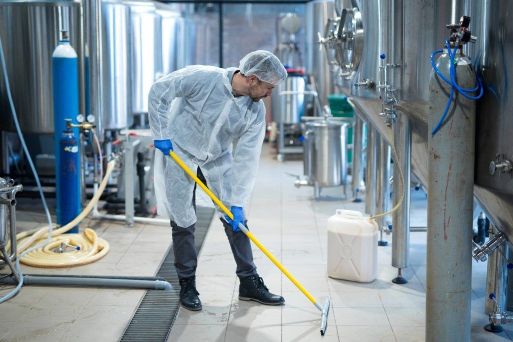 Ein Arbeiter in Schutzkleidung reinigt mit einem gelben Mopp einen nassen Fabrikboden in einer Industrieanlage aus Edelstahl.