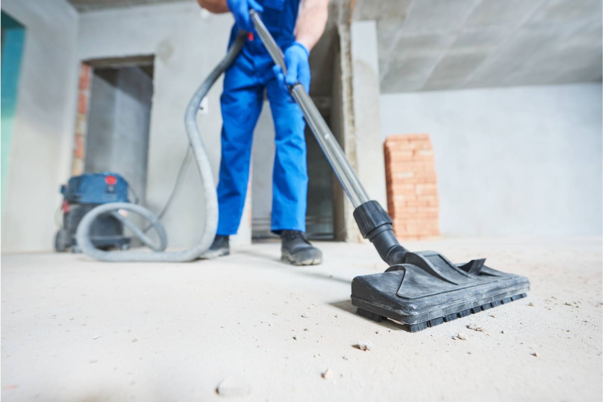 Ein Arbeiter in blauem Overall saugt Staub von einem Betonboden auf einer Baustelle ab.