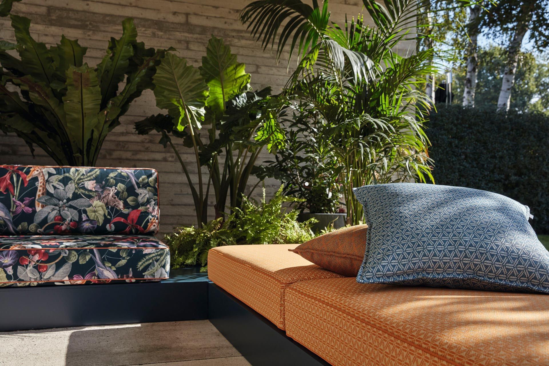 Ein Sofa mit Kissen steht vor einer Backsteinmauer, umgeben von Pflanzen.