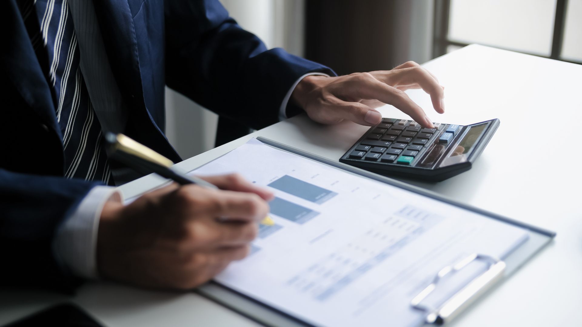 Une personne en costume effectue des calculs sur une calculatrice tout en consultant un bloc-notes contenant des graphiques et des tableaux.