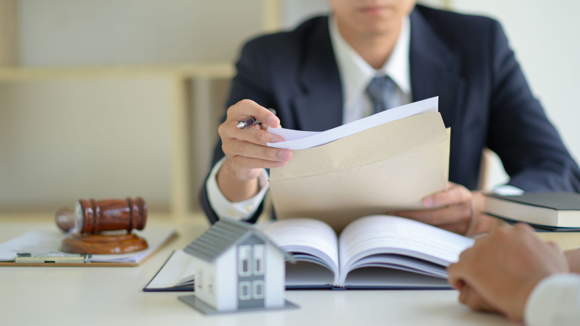 Un avocat en costume examine des documents avec un client à un bureau, un marteau et une maquette de maison sont présents.