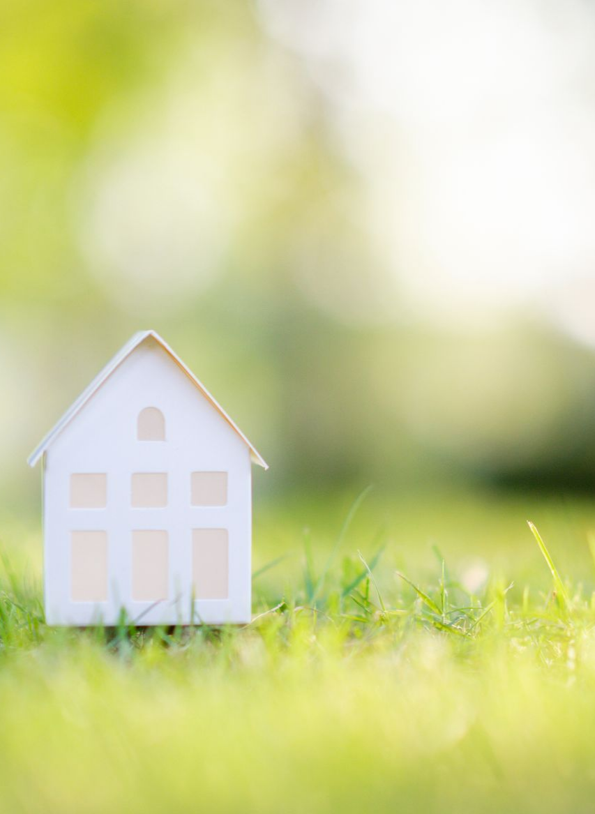 Maison miniature blanche sur herbe verte, arrière-plan flou de verdure et de lumière du soleil.