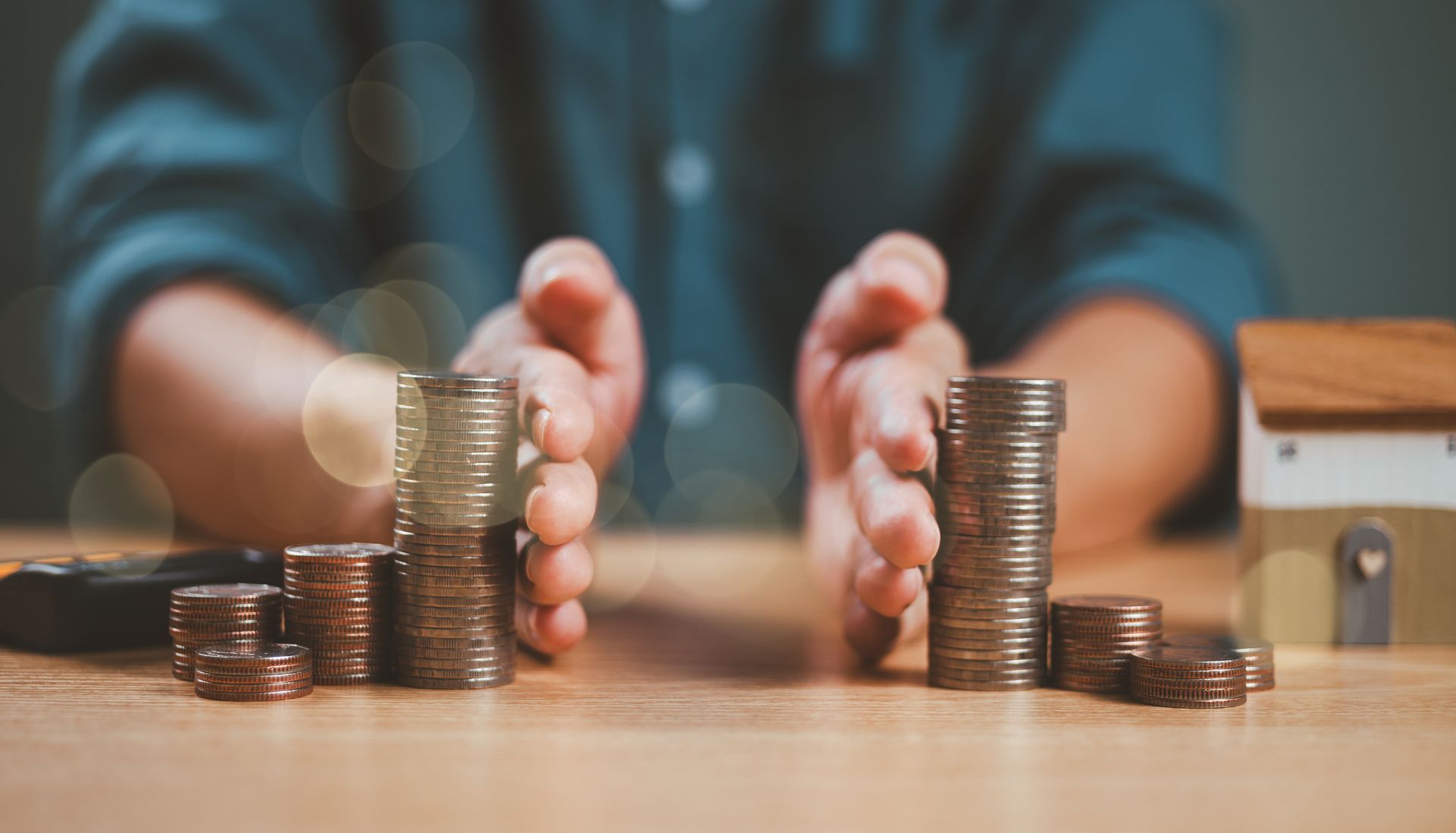 Des mains écartent des piles de pièces de monnaie posées sur une surface en bois, avec une maison de poupée et une calculatrice à proximité.