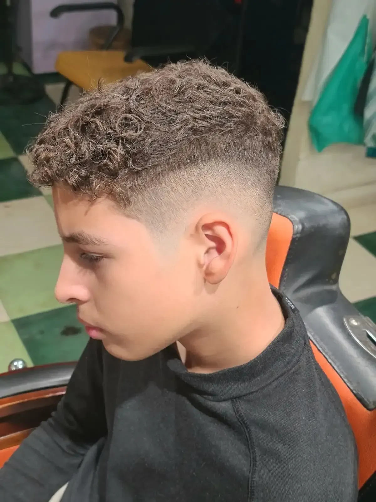 Corte de pelo de perfil con la parte superior rizada y los lados degradados, sentado en una silla de barbero.