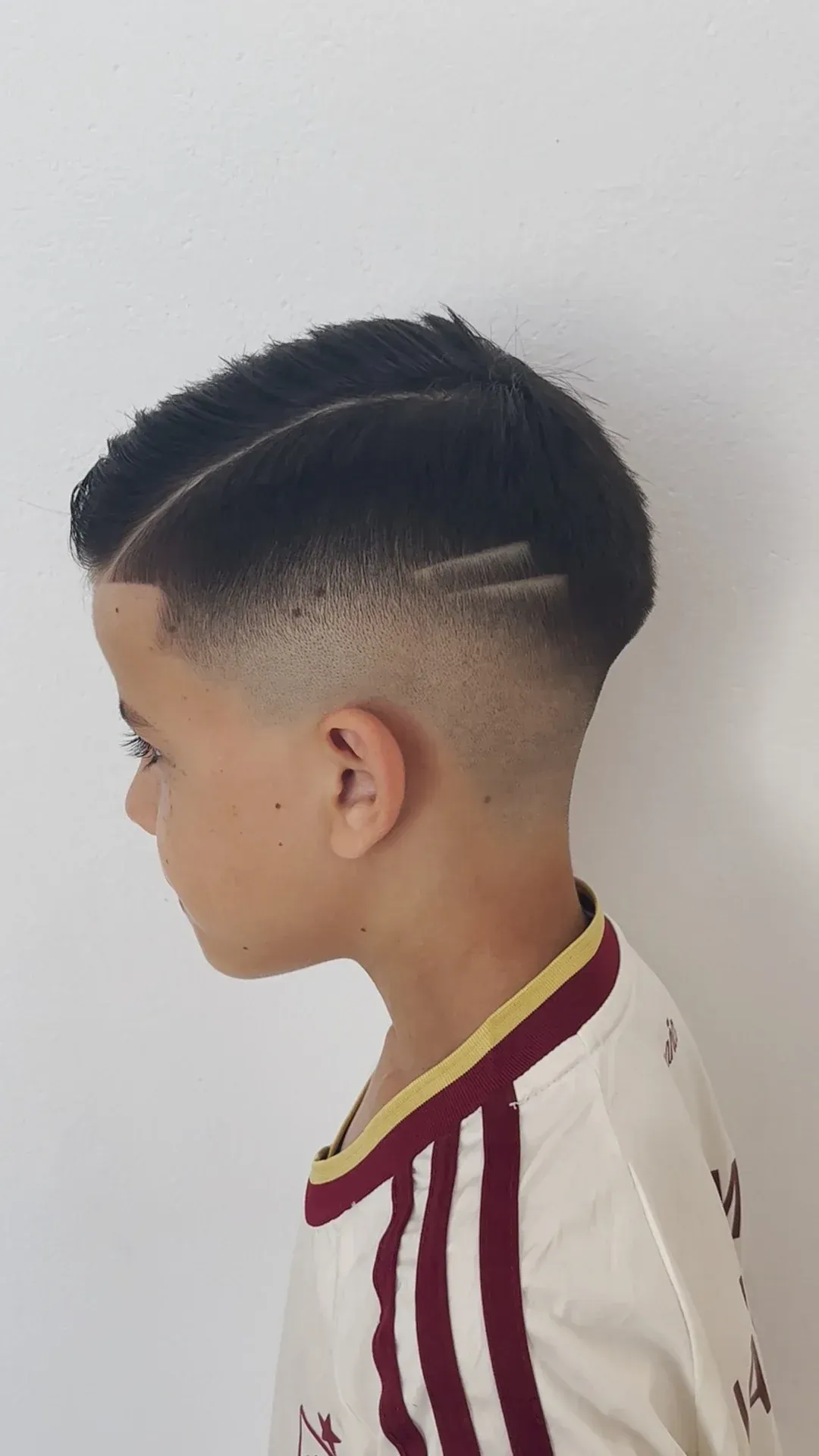 Perfil de un niño con un corte de pelo degradado alto que lleva una camisa blanca con rayas granates.