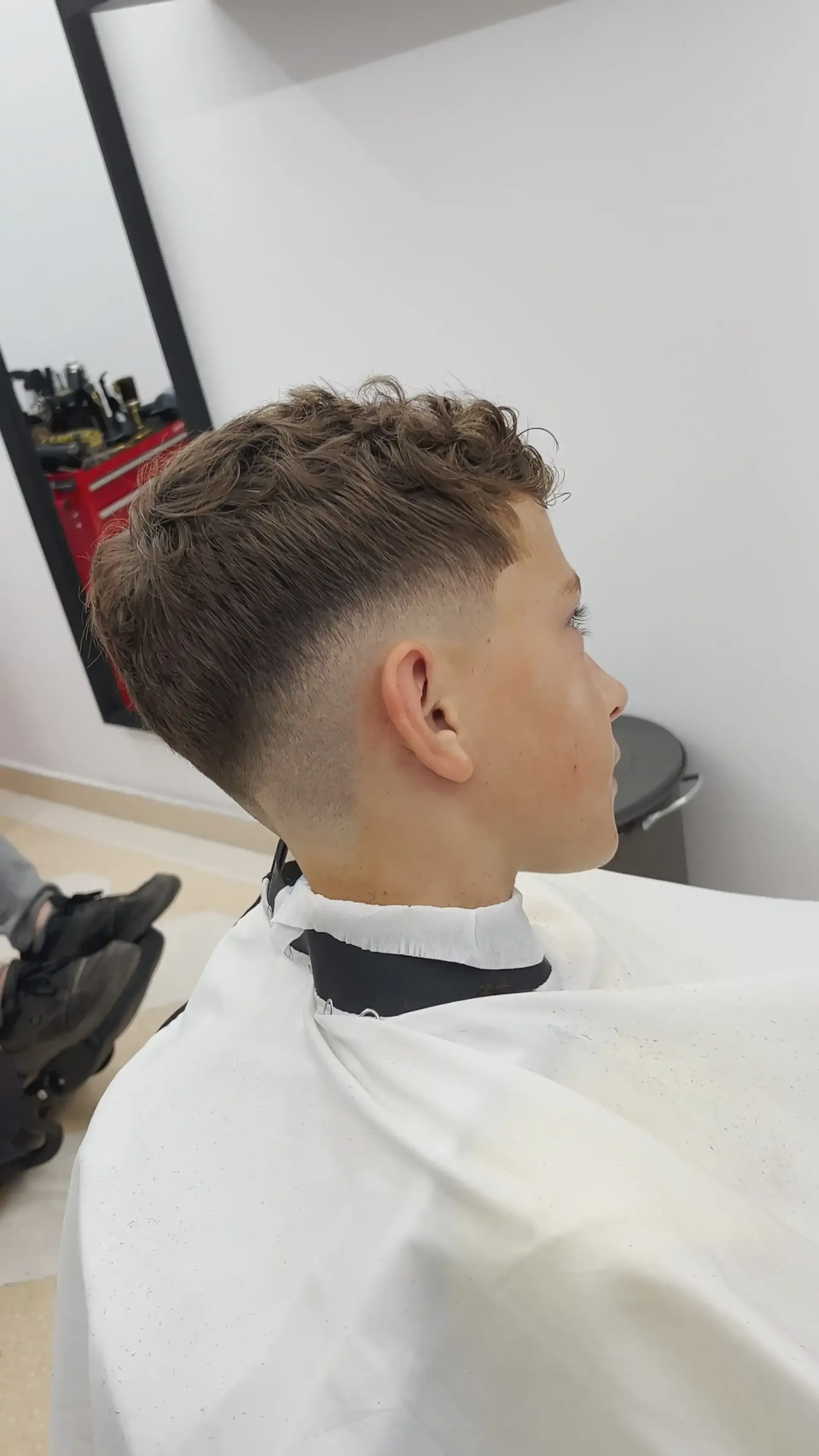 Perfil lateral de un corte de pelo corto y rizado con degradado en una barbería, con una capa blanca alrededor del cliente.