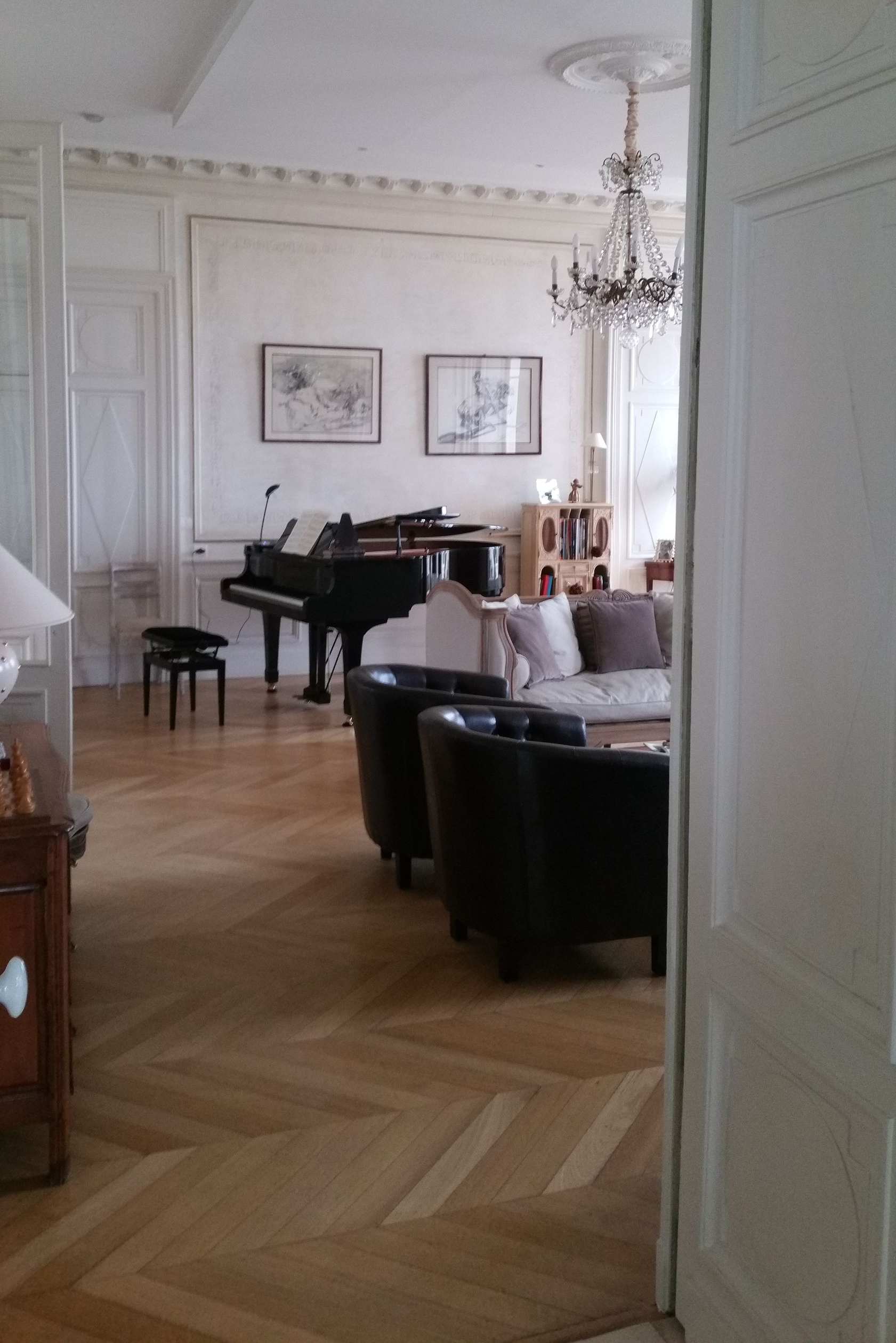 Salon avec piano à queue, coin salon et lustre. Parquet, murs blancs.