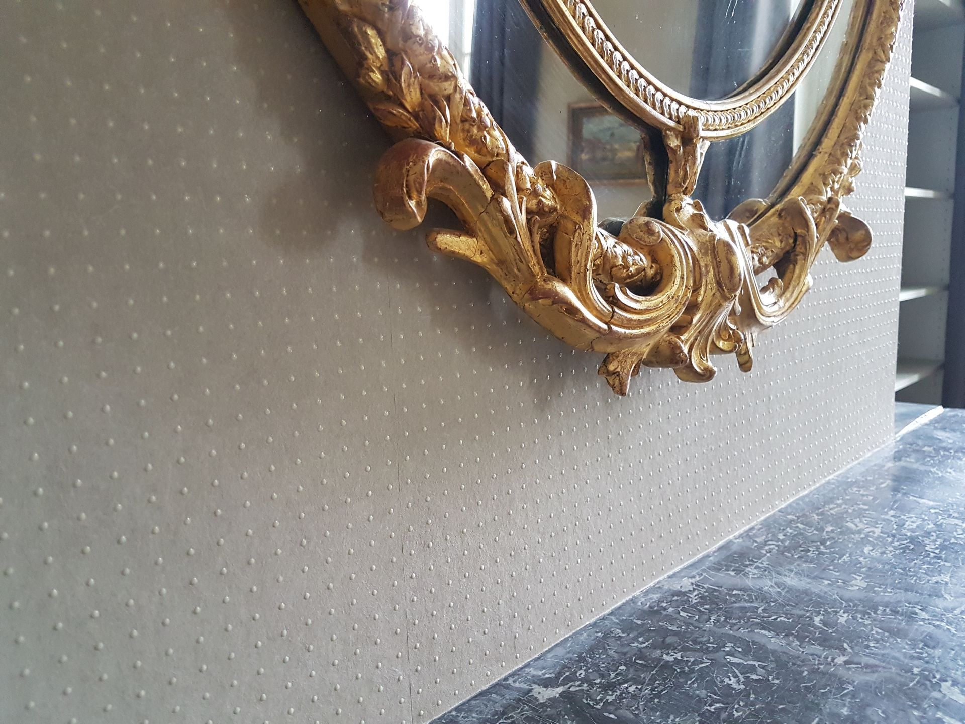 Miroir ovale orné d'un cadre doré, sur un mur texturé gris clair, posé sur un comptoir foncé.