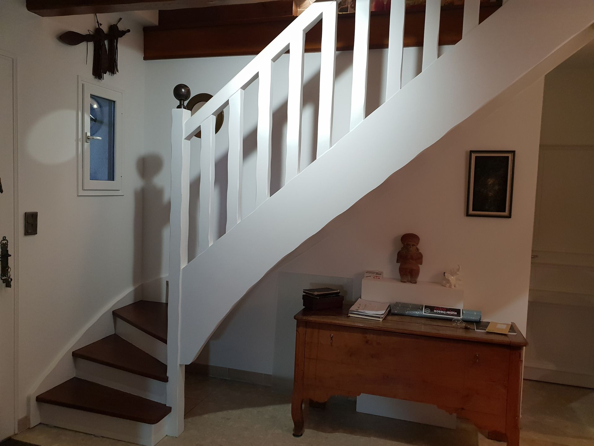 Escalier blanc aux marches en bois menant à un palier. Coffre en bois en dessous, petite fenêtre sur le côté.
