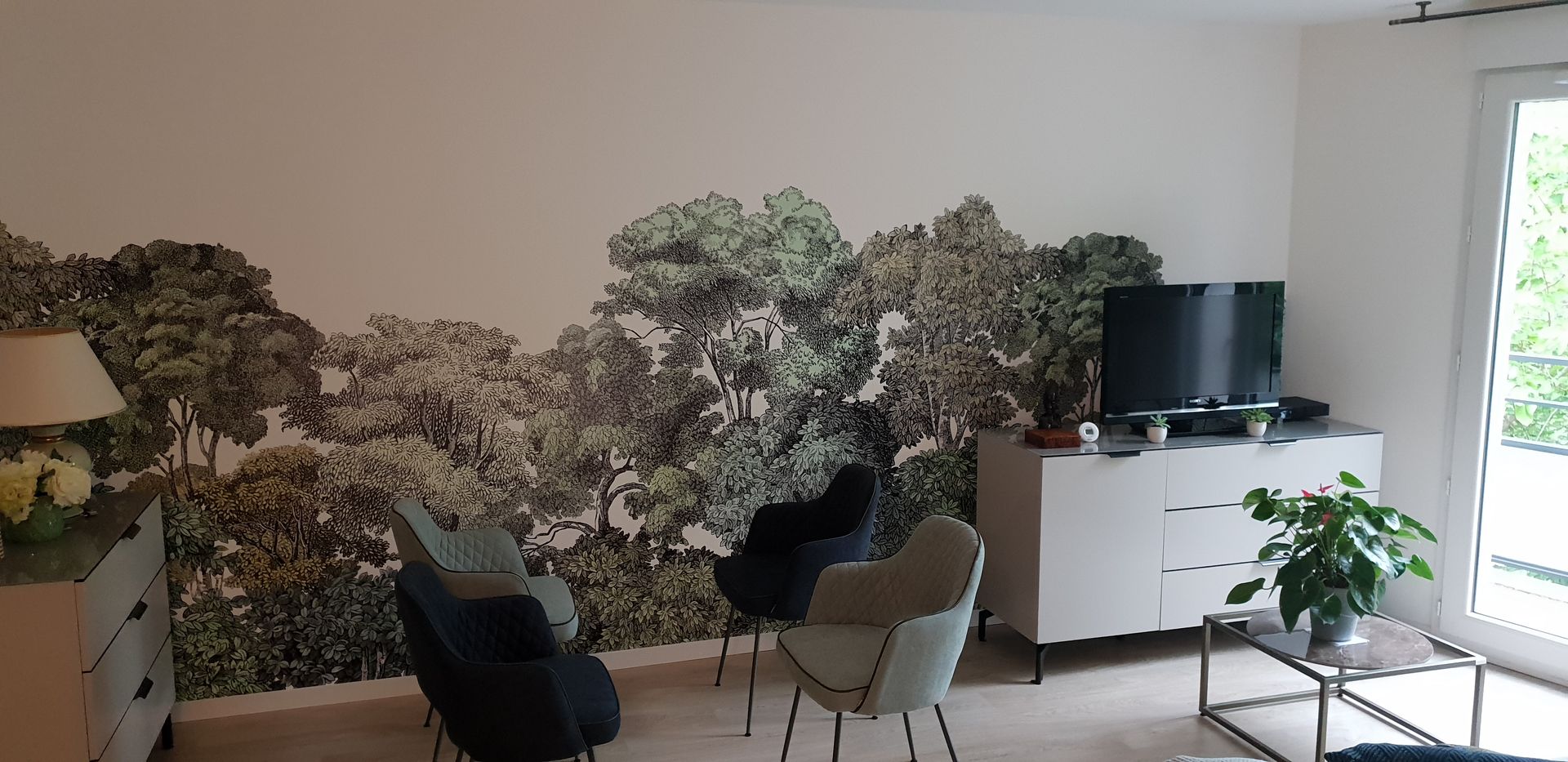 Salon avec fresque murale représentant une forêt, meuble blanc, télévision, deux fauteuils et une plante.