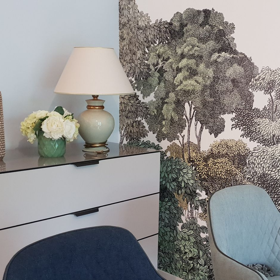 Fauteuil rose, petite table verte avec des vases, sur un papier peint bleu sarcelle foncé à motifs dorés.