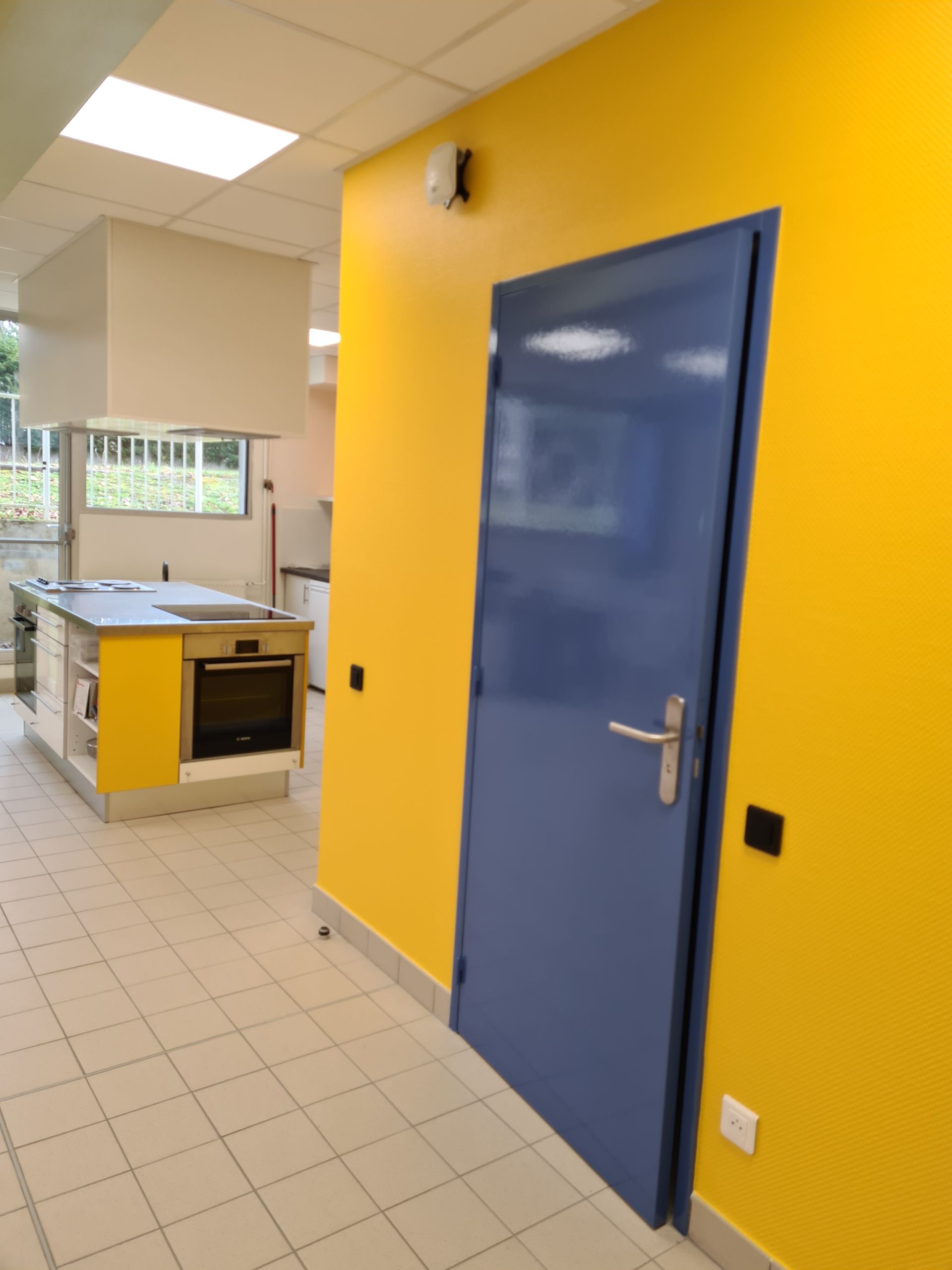Porte bleue dans une pièce aux murs jaunes, avec une cuisine visible en arrière-plan.