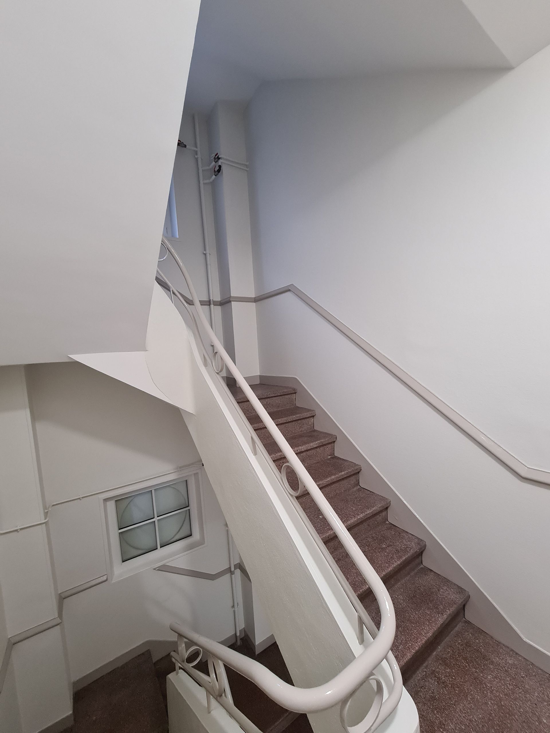 Cage d'escalier aux murs blancs, marches et rampes en granit. Fenêtre visible au niveau inférieur.