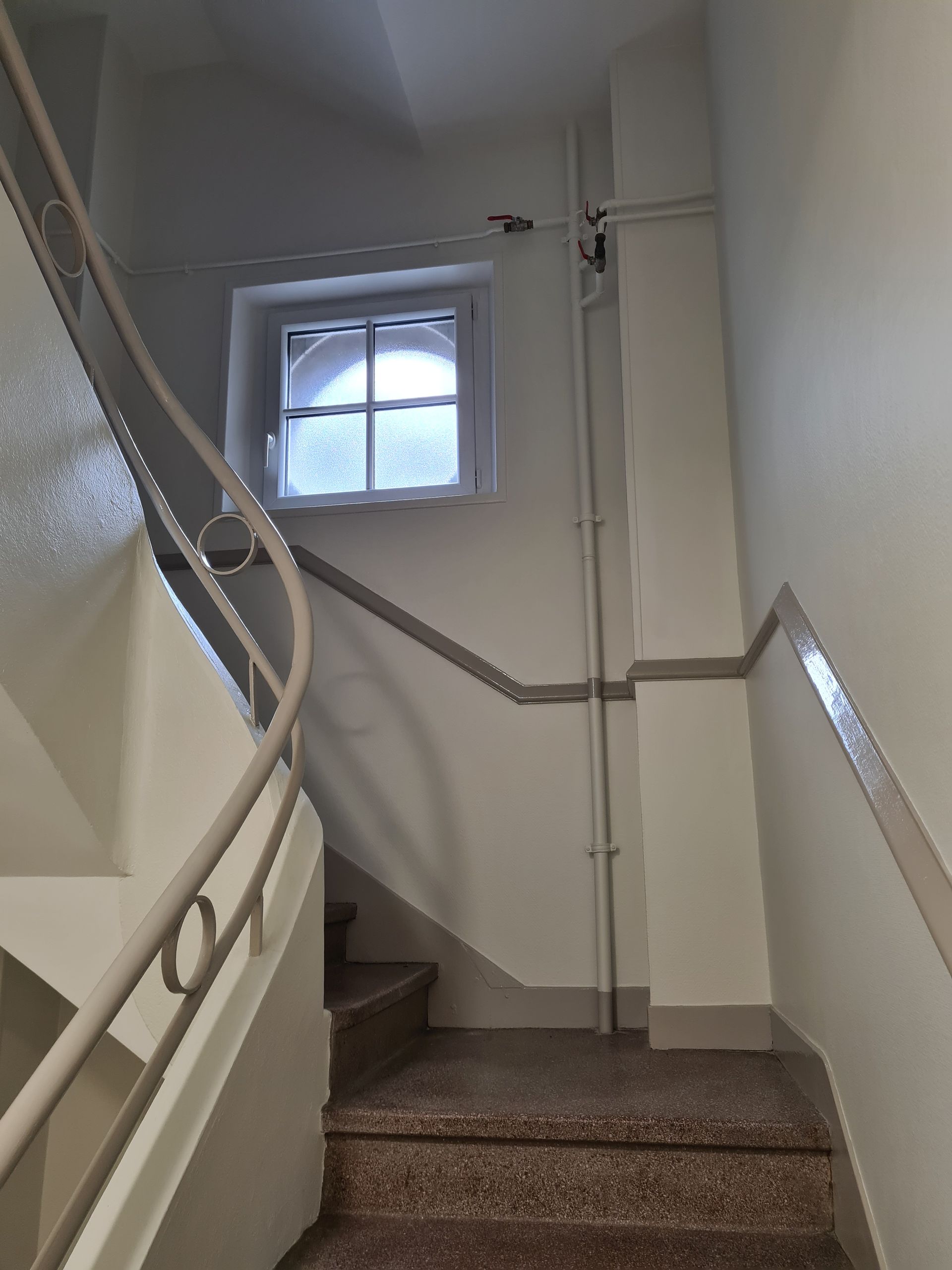 Escalier aux marches de marbre, murs blancs et petite fenêtre cintrée. Une rampe décorative s'incurve vers le haut.