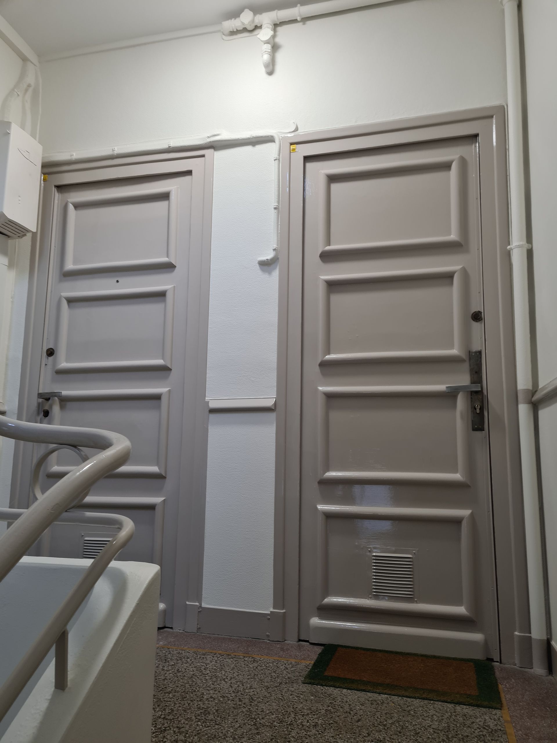 Deux portes grises dans un couloir, l'une avec une petite fenêtre, à côté d'une cage d'escalier avec une rampe.