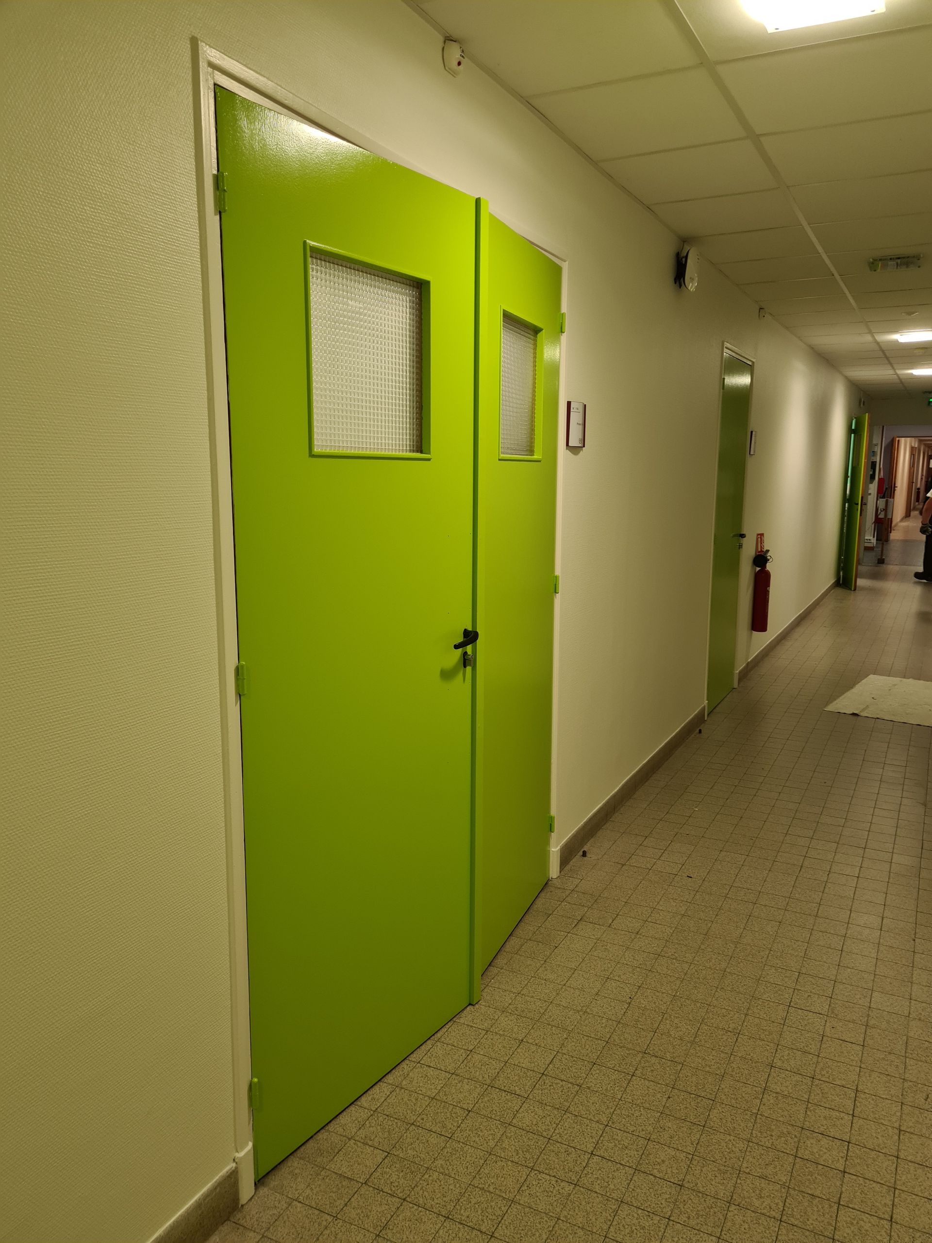 Double porte verte avec petites fenêtres dans un couloir blanc.