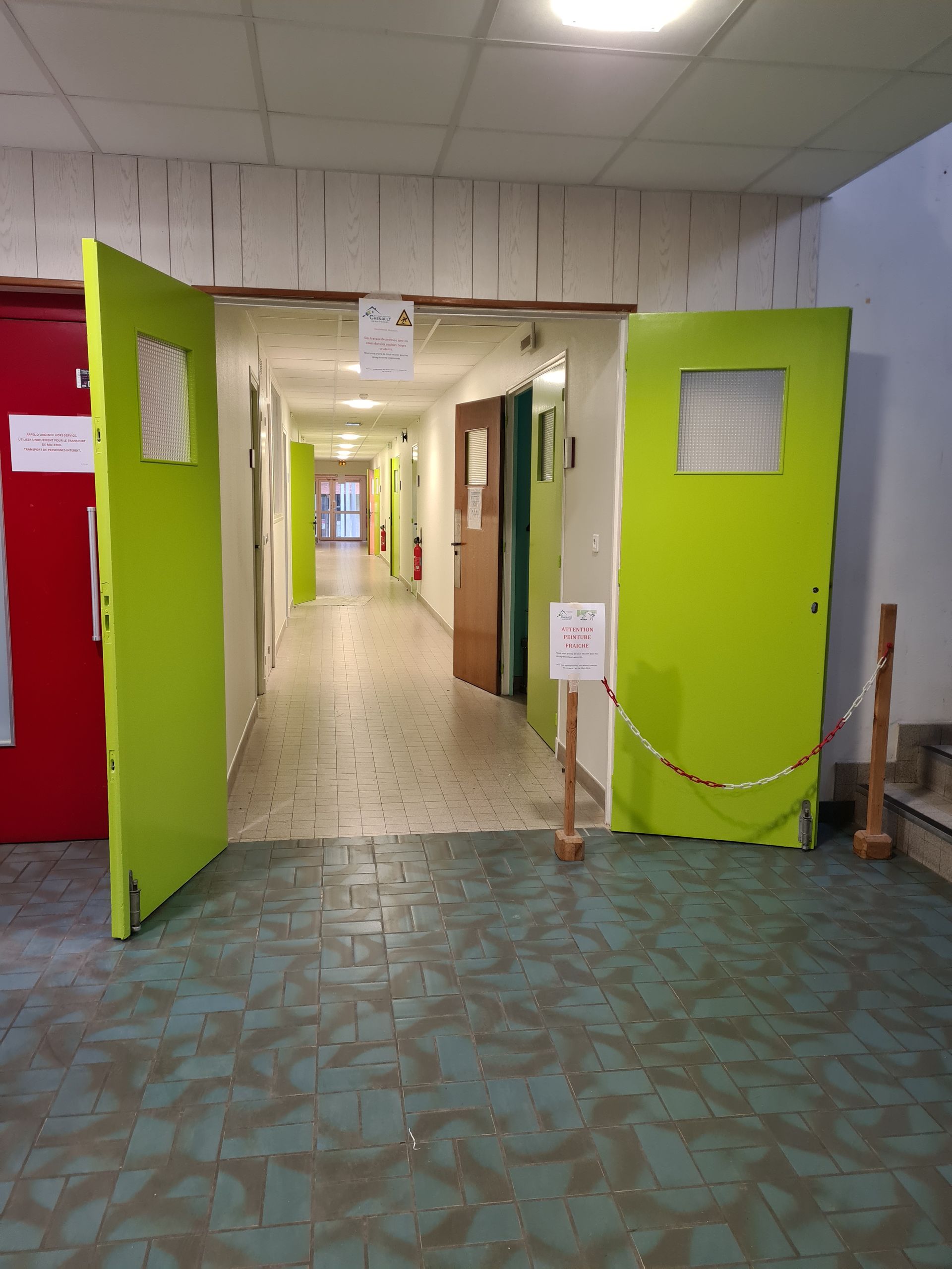 Couloir avec portes vertes et rouges, ouverture sur un long couloir. Sol carrelé.