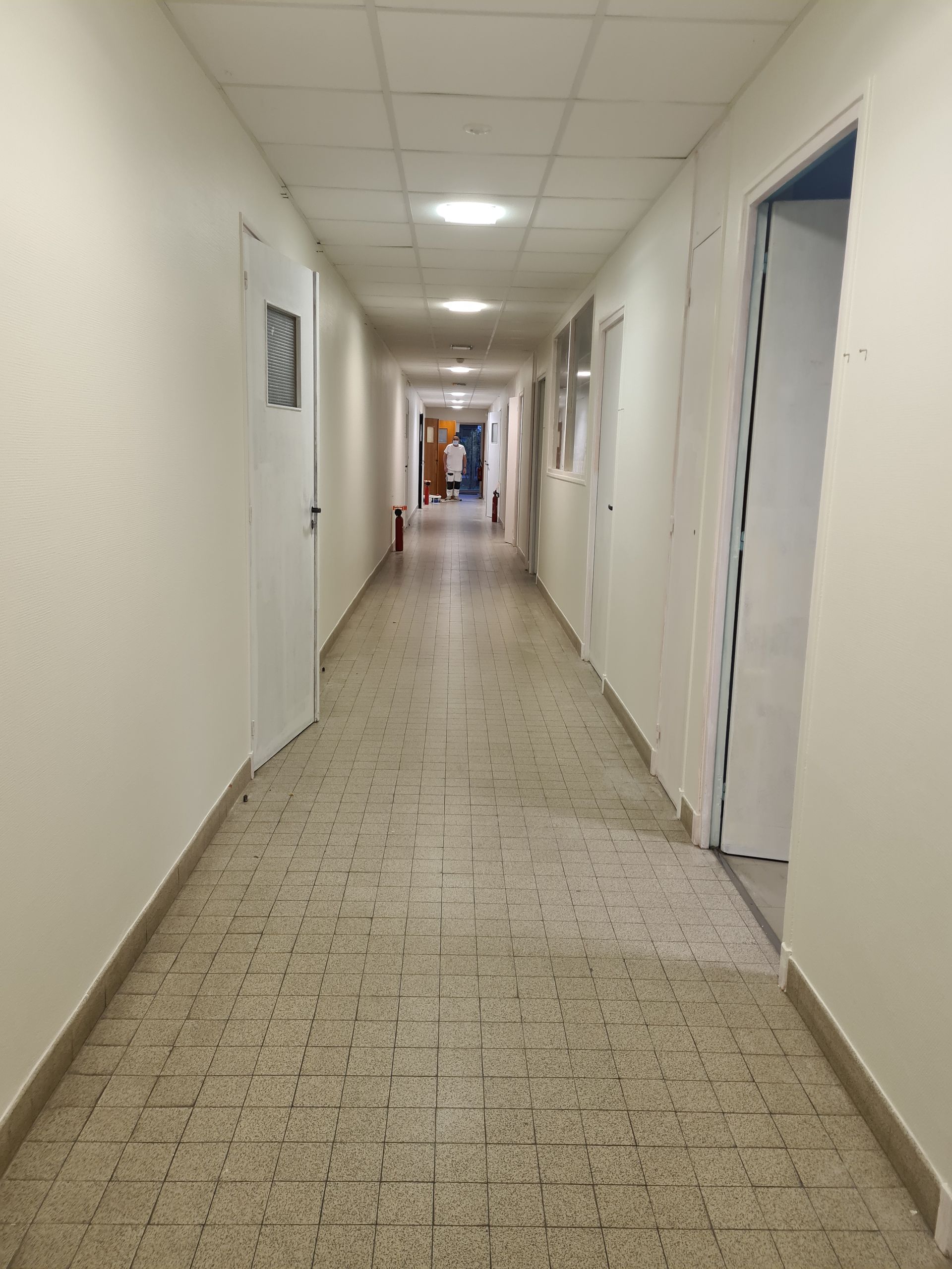 Long couloir étroit aux murs blancs et au sol carrelé. Plusieurs portes bordent les deux côtés.