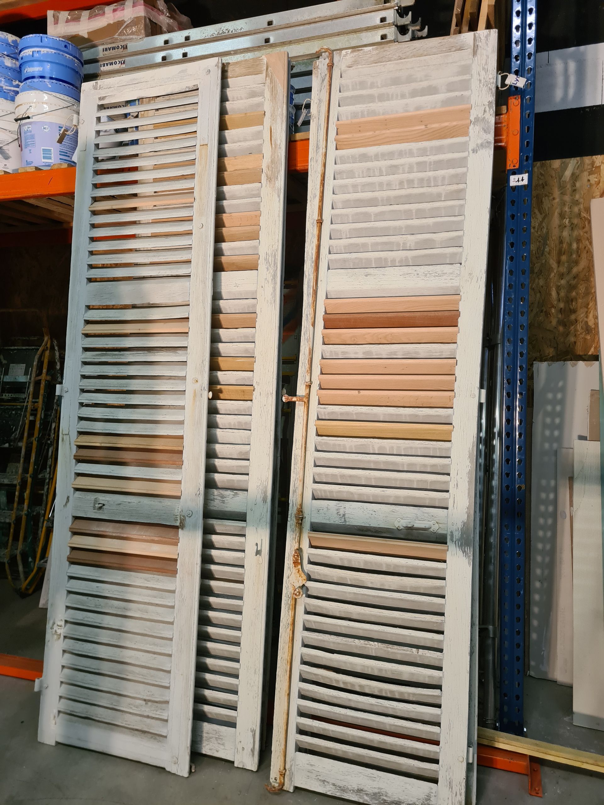 Deux volets en bois peints en blanc, patinés par le temps et à lattes horizontales, appuyés contre une étagère de rangement.