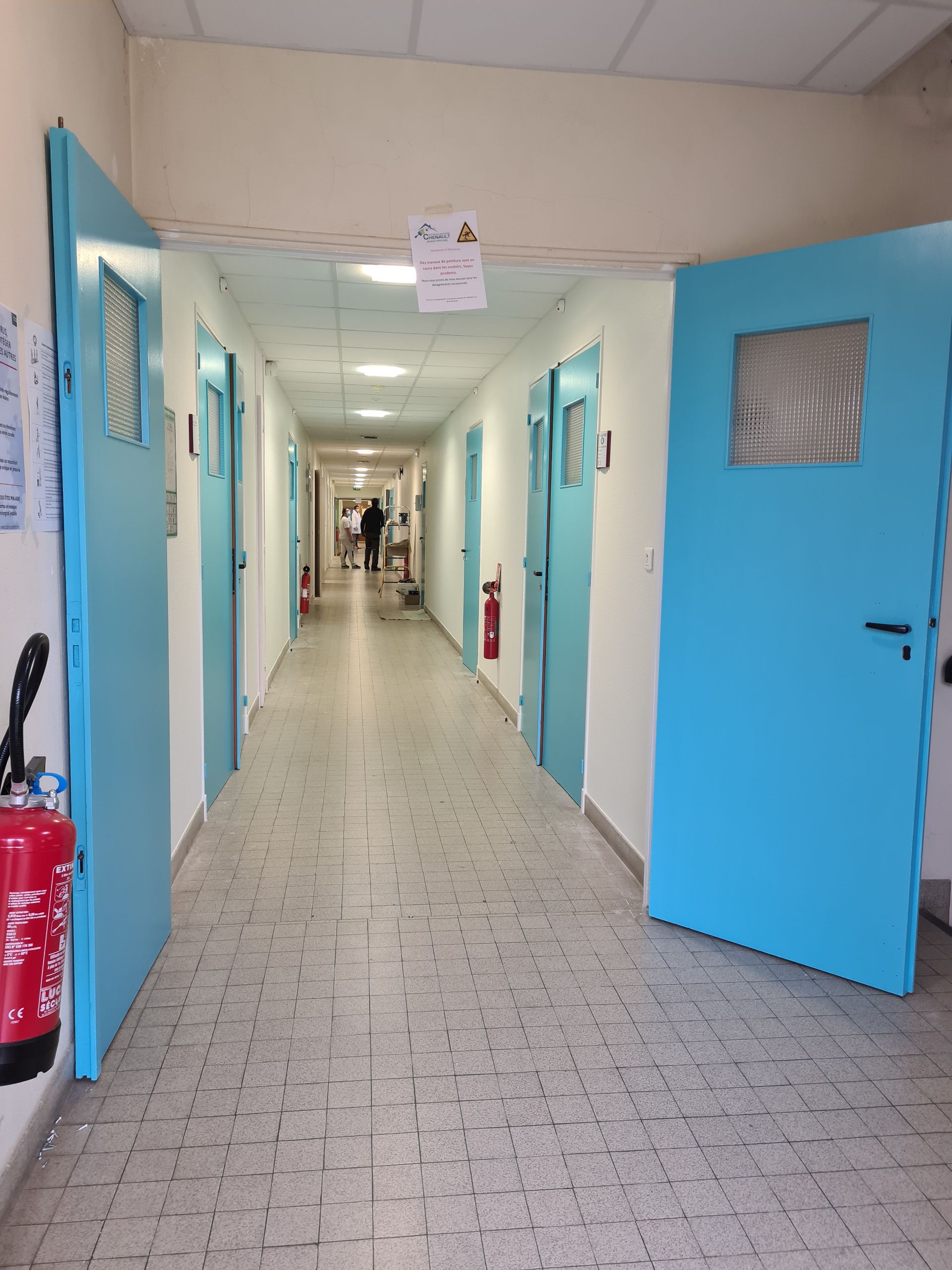 Long couloir d'hôpital avec des portes bleues et une personne qui marche vers le fond.