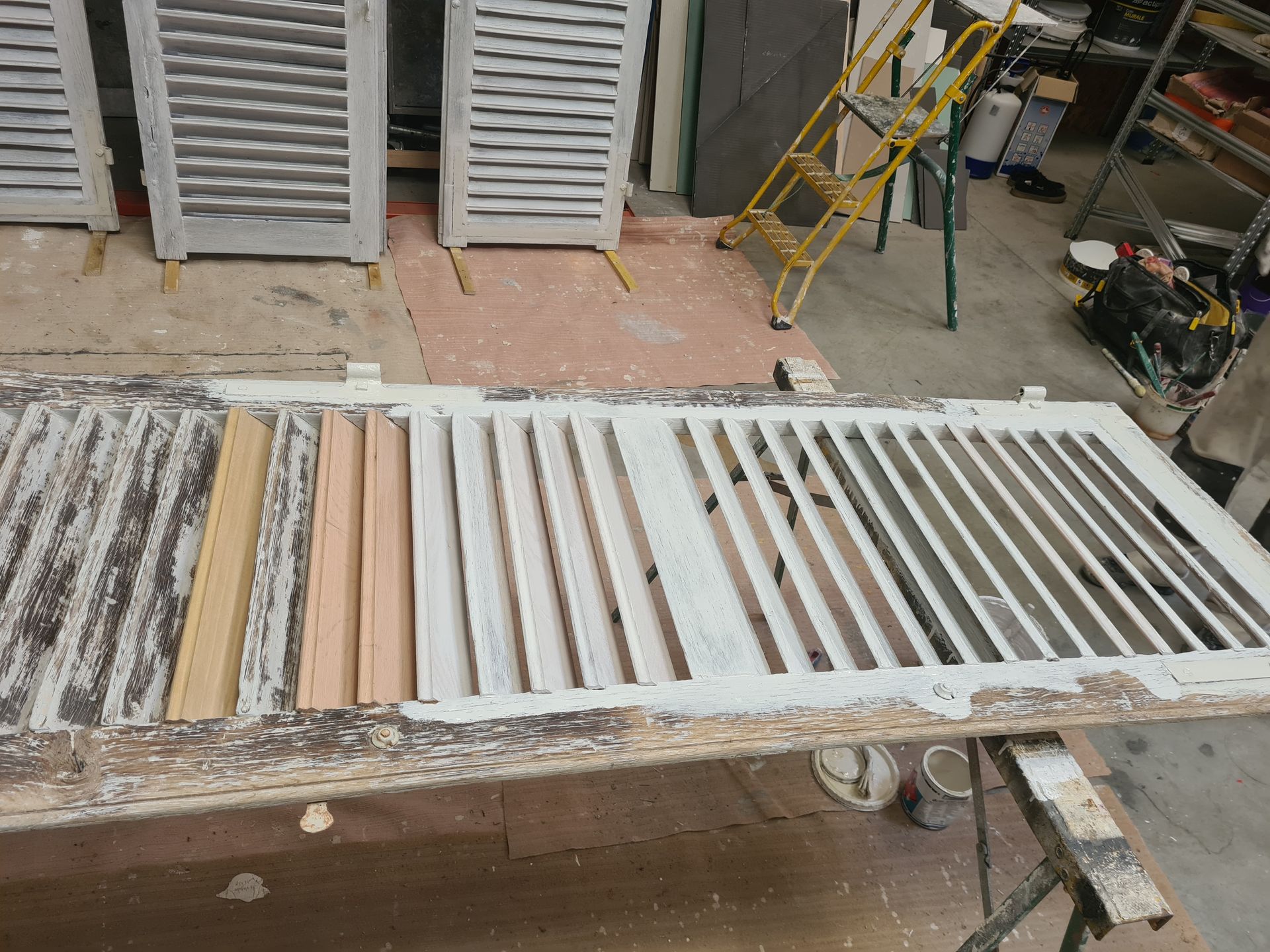 Un volet blanc partiellement démonté, avec des lamelles de rechange, sur un établi dans un atelier.