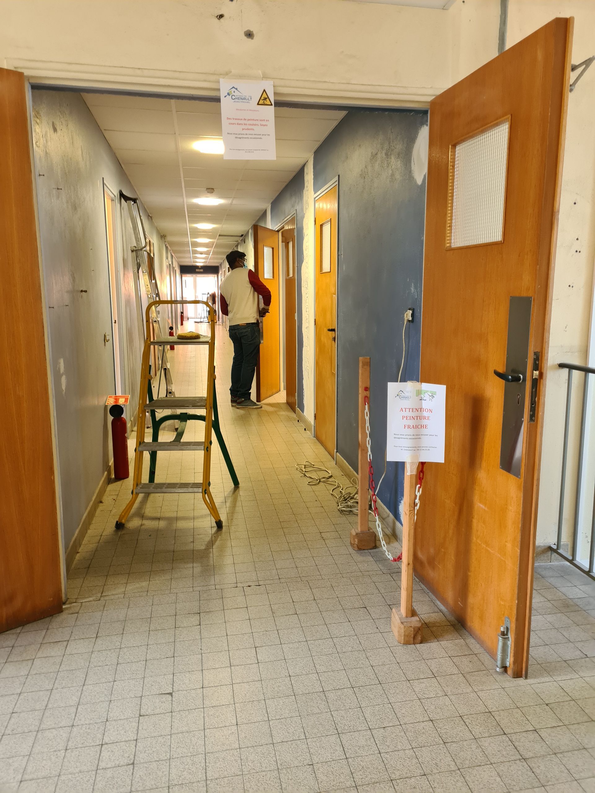 Couloir avec portes ouvertes, une personne s'éloigne. Une échelle et des panneaux de signalisation sont présents.