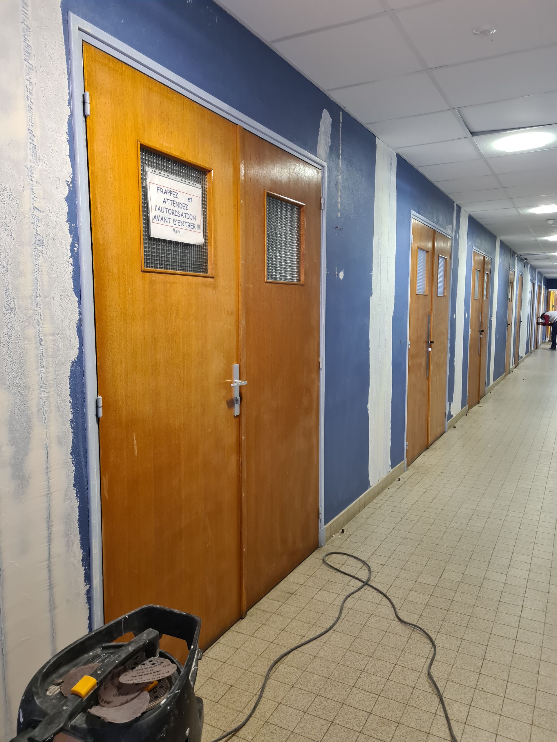 Couloir aux murs bleus, portes marron et sol carrelé. Un pot de peinture est posé au sol près de la première porte.